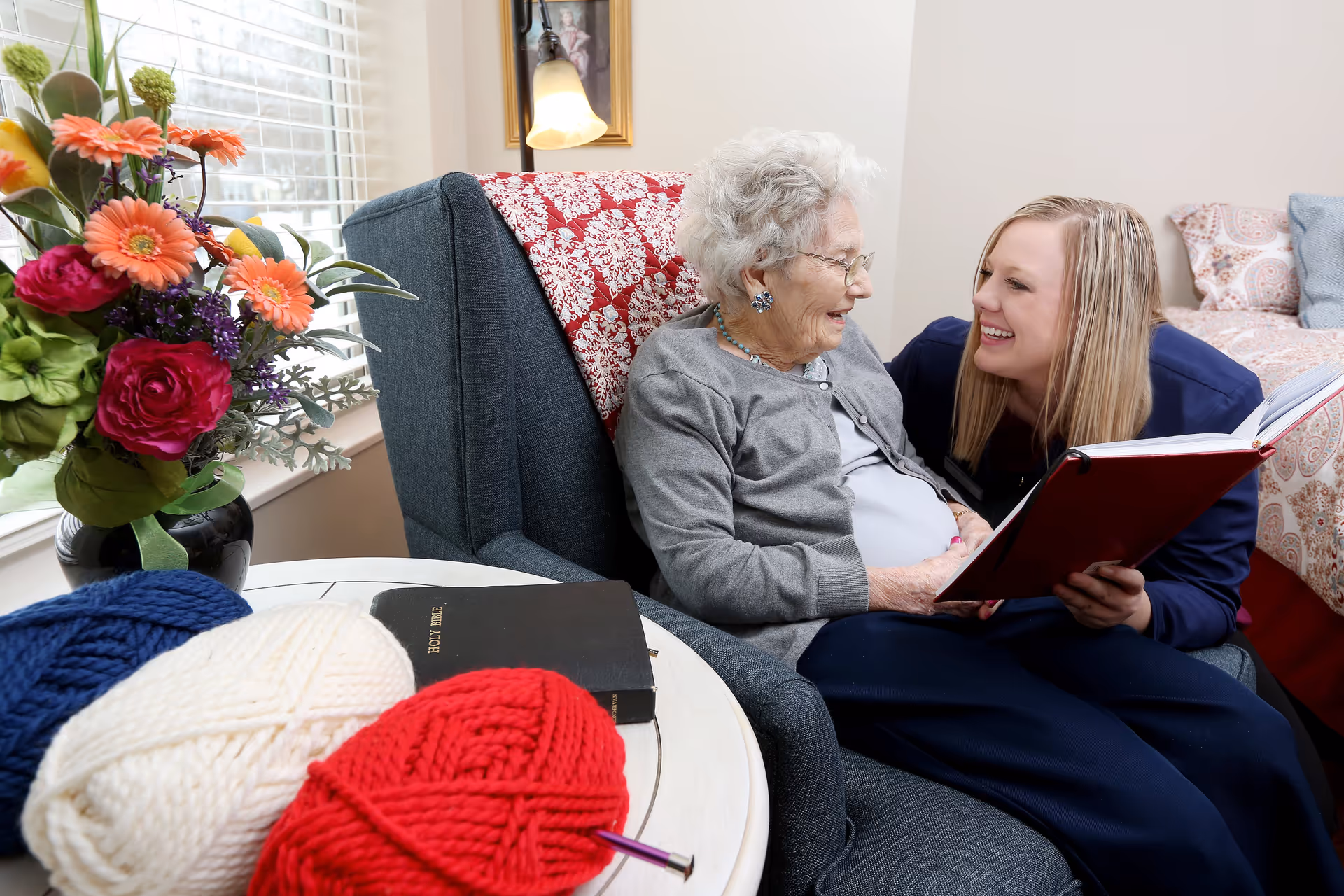 An elderly woman sitting in a blue armchair with a red and white patterned blanket behind her, smiling and looking at a younger woman who is holding an open red book and smiling back. A table next to the armchair holds a vase with colorful flowers, three large balls of yarn in blue, white, and red, and a black Holy Bible. A bed with patterned pillows is visible in the background.