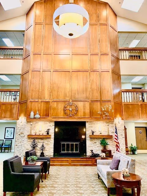 A spacious and well-lit living room area with a high wooden paneled wall featuring a large clock above a black fireplace. The room has comfortable seating including two dark armchairs and a light-colored sofa with cushions. There are decorative plants, sculptures, and an American flag near the fireplace. The upper level has wooden railings overlooking the room, and a large modern circular light fixture hangs from the ceiling.