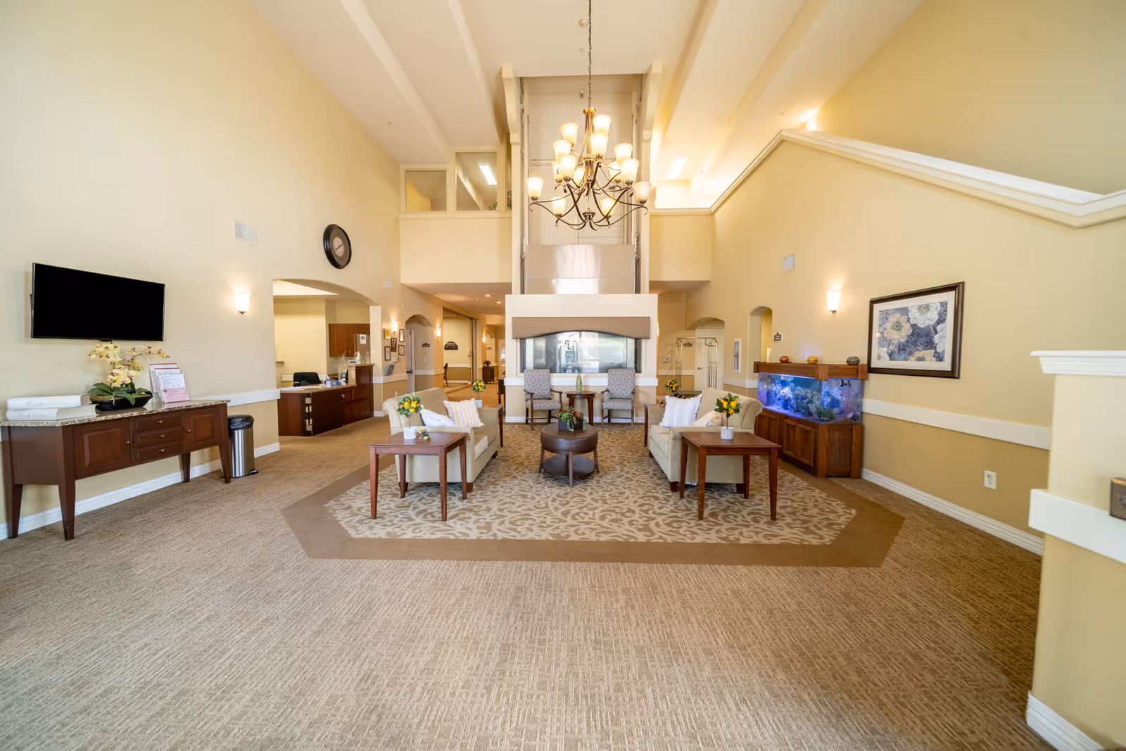 A spacious and well-lit living room area in a senior living facility with beige walls and carpet. The room features two beige sofas with pillows, two armchairs, a central coffee table, side tables with flower vases, a large chandelier hanging from the ceiling, a wall-mounted TV, a large fish tank, and a fireplace. There is a reception desk visible in the background.