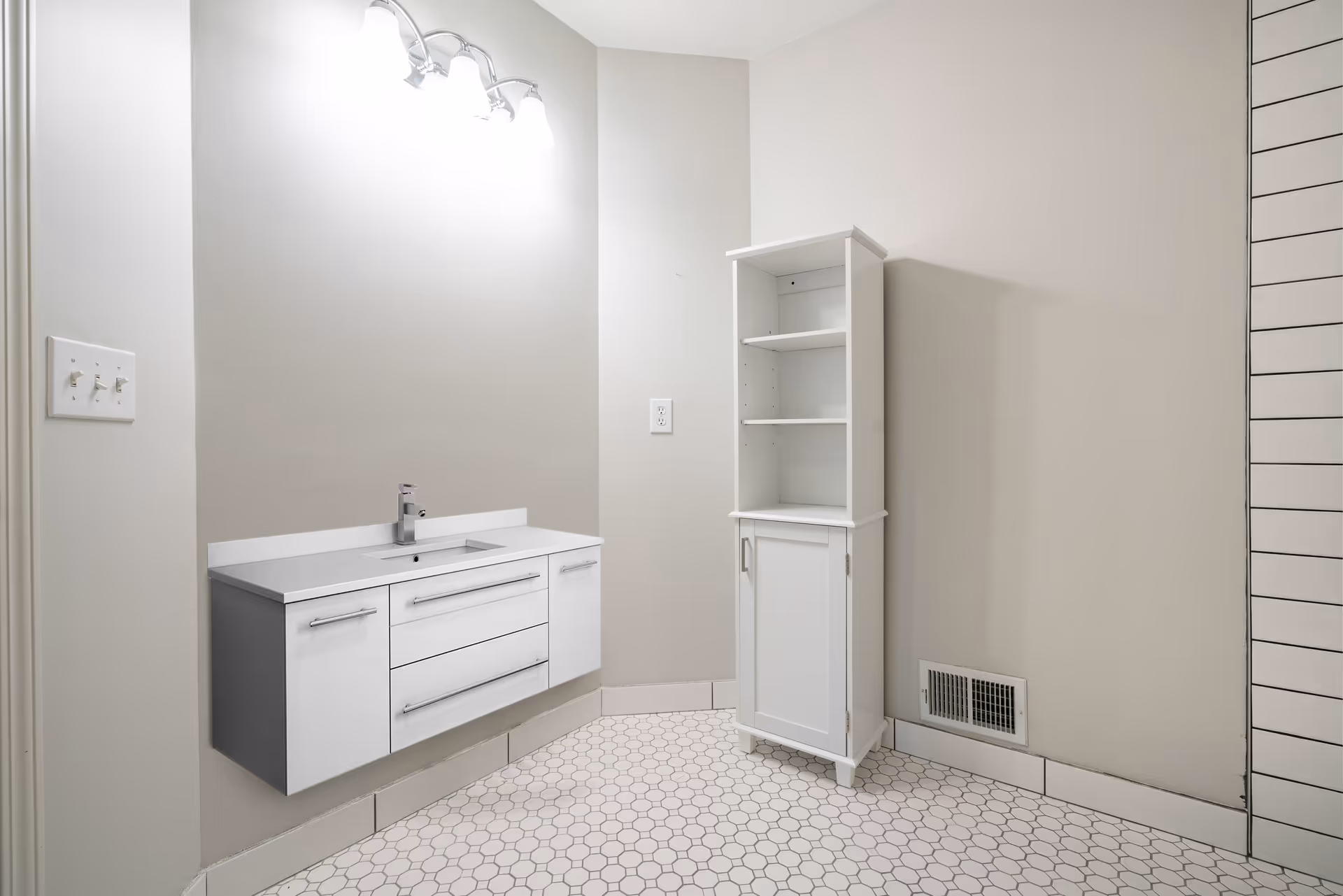 A modern bathroom with light gray walls and white hexagonal tile flooring. There is a floating white vanity with a built-in sink and a chrome faucet on the left wall, illuminated by a three-light fixture above. On the right side, there is a tall white cabinet with open shelves and a closed compartment at the bottom. A vent is visible near the floor on the right wall.