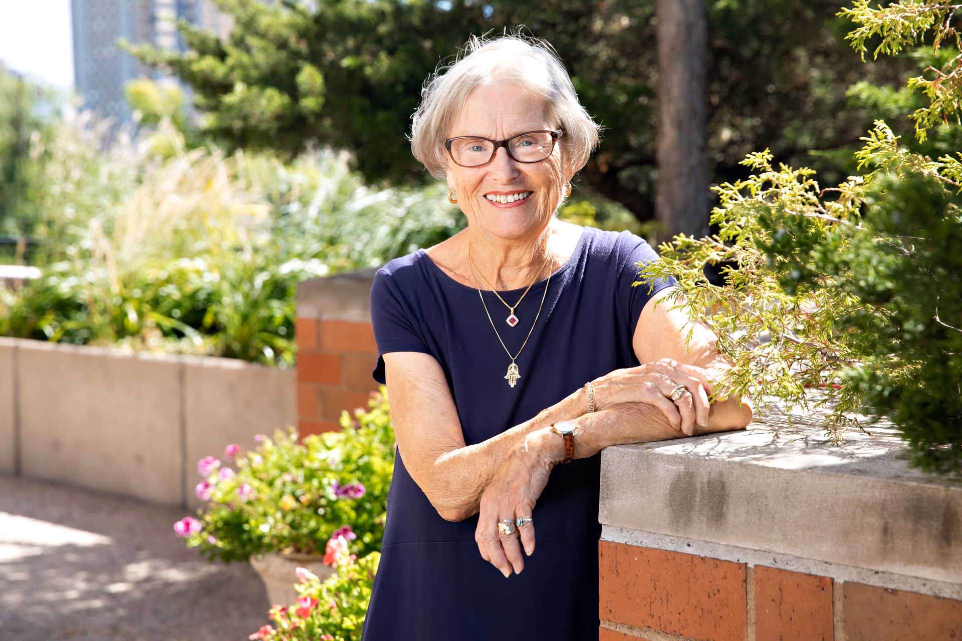 An elderly woman with short gray hair and glasses, wearing a navy blue dress and jewelry, smiling and leaning on a brick and concrete ledge in a garden area with green plants and flowers in the background.