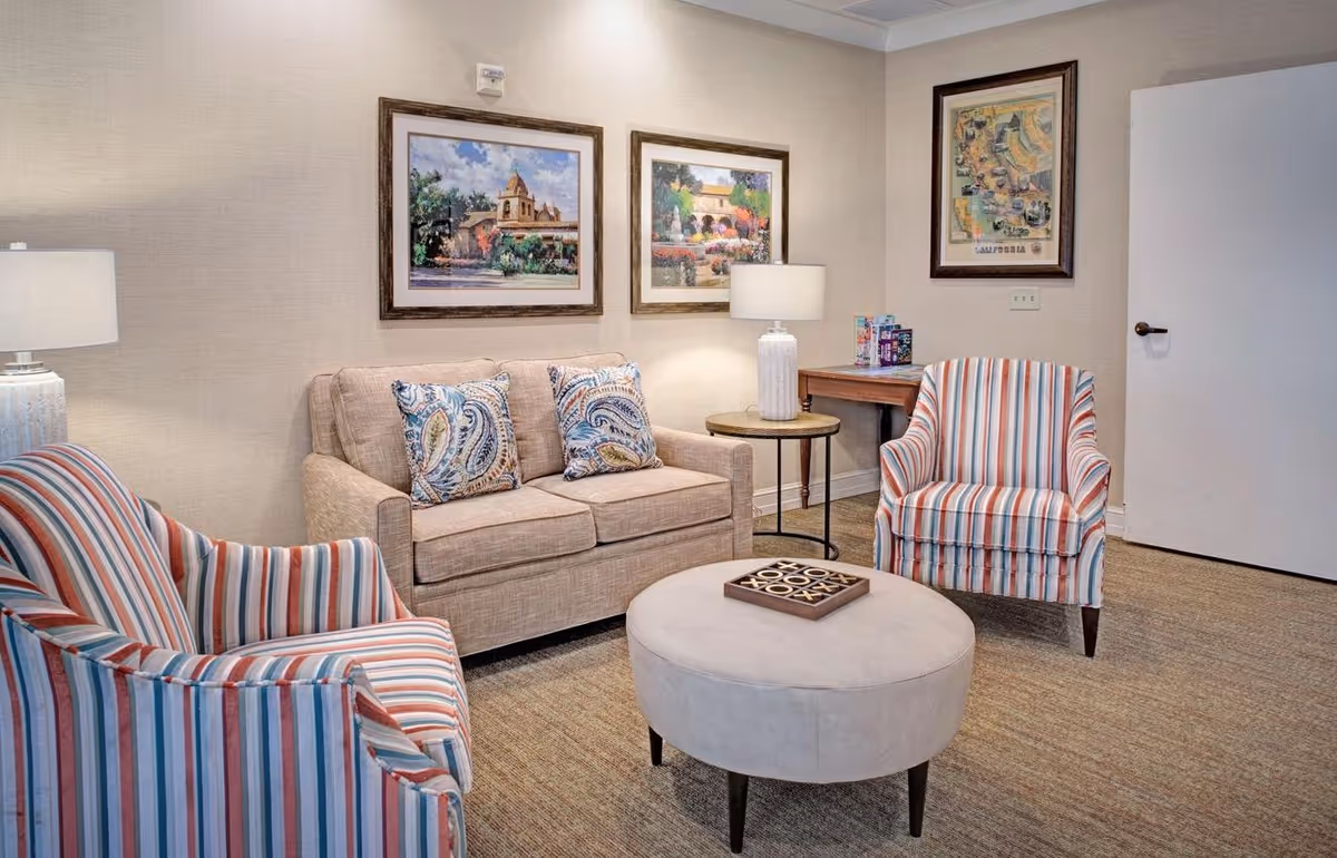 A cozy sitting room with a beige loveseat, two striped armchairs, a round ottoman, side tables with lamps, and framed artwork on the walls.