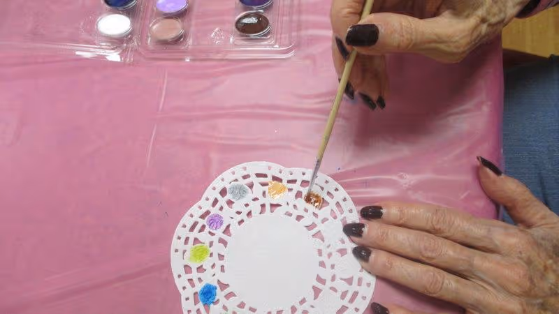 An elderly person with dark nail polish is painting a white paper doily with various colors of paint on a pink tablecloth. A paintbrush is held in one hand, applying orange paint to the doily, while the other hand steadies it. A palette with multiple paint colors is visible in the upper left corner.