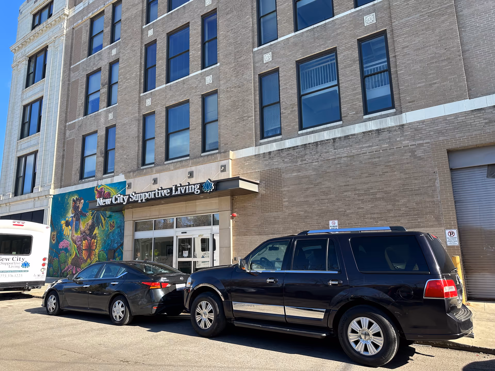 Front exterior of a multi-story brick building showing the entrance and sign 'New City Supportive Living', a colorful mural to the left, and parked cars in front.