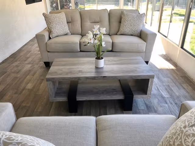 Bright sunlit living room with beige sofas and a wooden coffee table holding a potted white orchid near large windows.