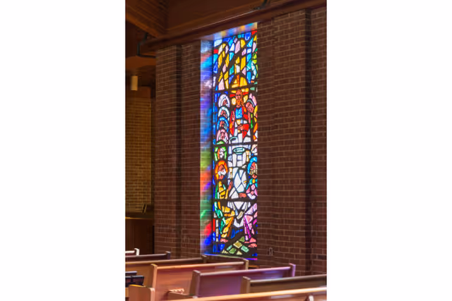 Interior view of a chapel or worship area with wooden pews and a tall, narrow stained glass window featuring colorful religious imagery set in a brick wall.