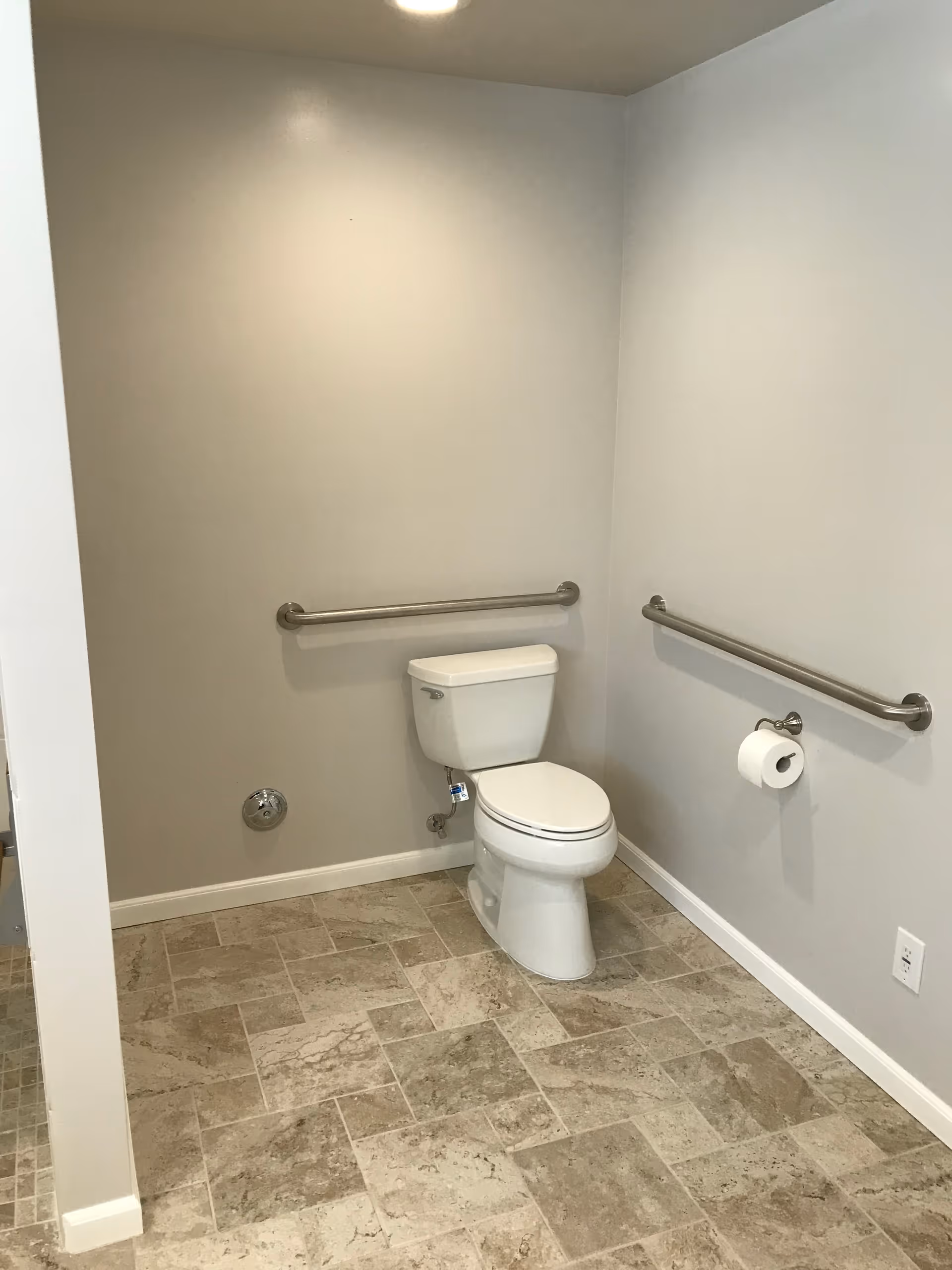 Accessible restroom with a toilet, grab bars on two walls, a toilet paper holder, and tiled floor.