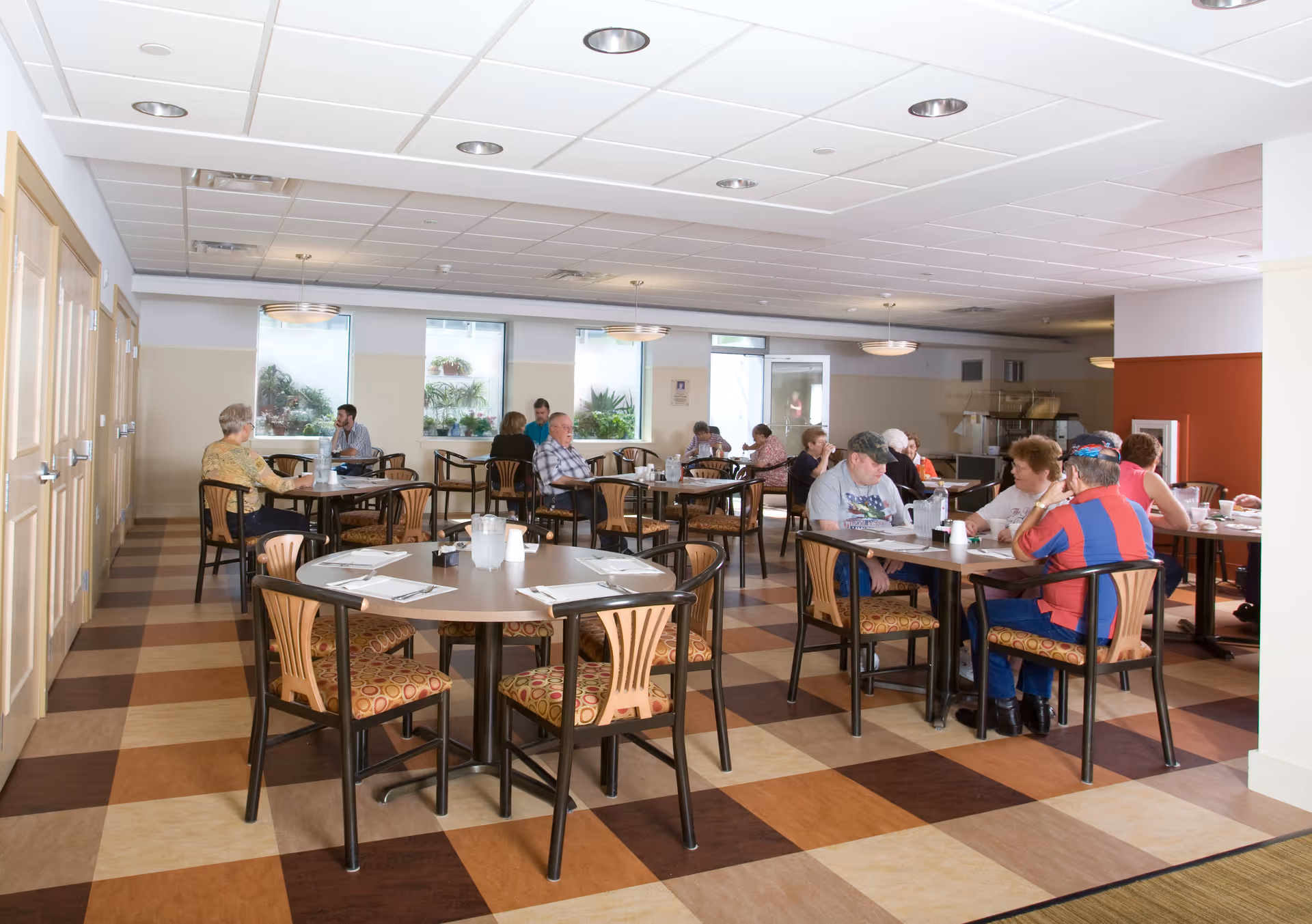 A dining room in a senior living facility with several round tables and chairs. Multiple elderly people are seated at the tables, eating and conversing. The room has large windows with plants outside, a checkered floor pattern, and ceiling lights.