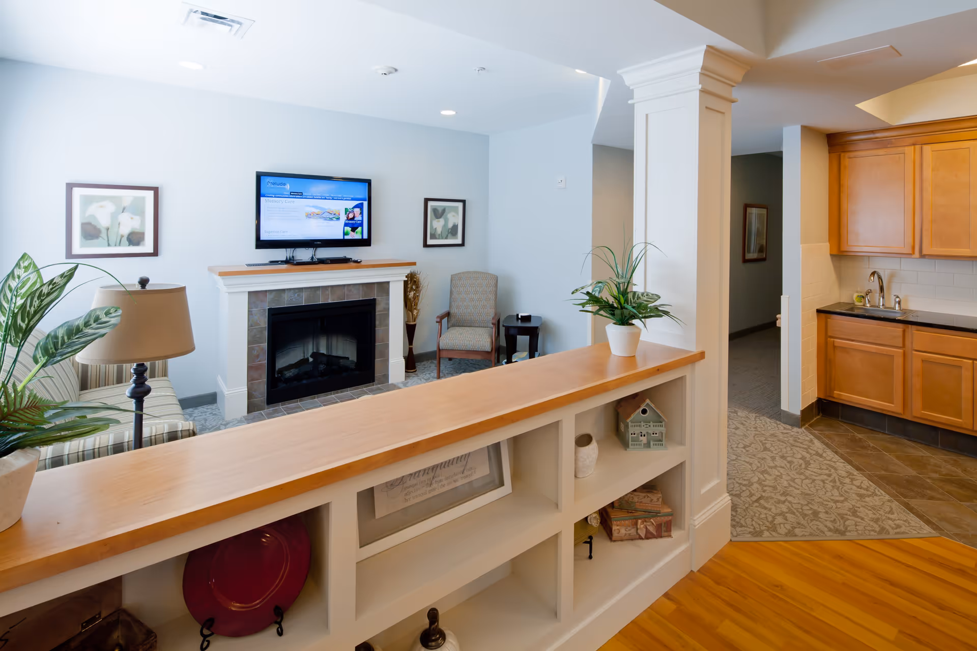 A cozy living room area in Prelude Homes & Services featuring a fireplace with a mounted flat-screen TV above it, two framed floral artworks on the wall, a patterned armchair next to a small side table, and a striped sofa partially visible on the left. In the foreground, there is a wooden countertop with built-in shelves holding decorative items and plants. To the right, there is a kitchenette area with wooden cabinets, a sink, and tiled flooring.