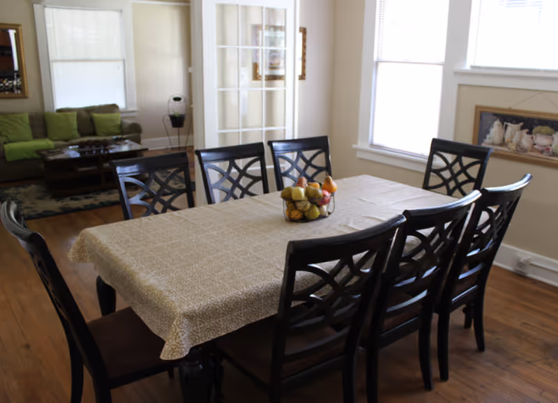 Dining room with a rectangular table covered by a patterned tablecloth surrounded by eight dark wooden chairs and a fruit bowl, with a living area visible in the background.