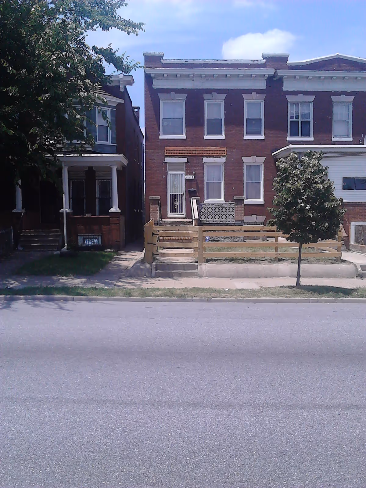 Front view of a two-story brick rowhouse with a small fenced front yard, sidewalk, and a tree.