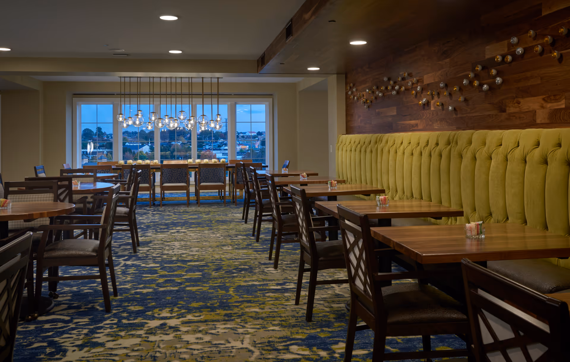 Dining room with wooden tables and chairs, a long green tufted banquette along a wood-paneled wall, and pendant lights by large windows.