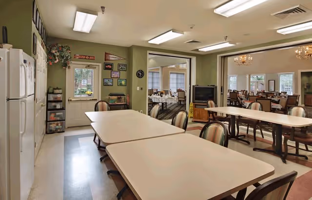 Interior view of a senior living facility room with several tables and chairs arranged for group activities or dining. The walls are painted green, and there is a refrigerator and storage drawers on the left side. A doorway leads to a dining room with multiple tables set with white tablecloths and chairs. The room has fluorescent ceiling lights and a television on a wooden stand.