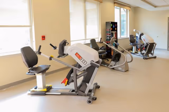 A bright exercise room with three seated fitness machines positioned near large windows with blinds. The room has light-colored walls and flooring, and a rack with colorful exercise balls is visible in the background.