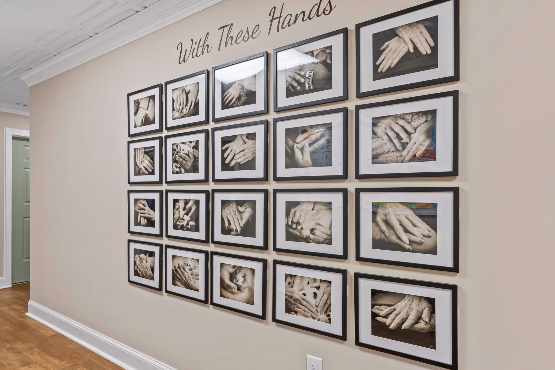 A beige wall with a grid of 20 framed black and white photographs showing close-ups of elderly hands in various poses. Above the frames, the words 'With These Hands' are written in cursive. The floor is wooden, and a green door is visible on the left side.