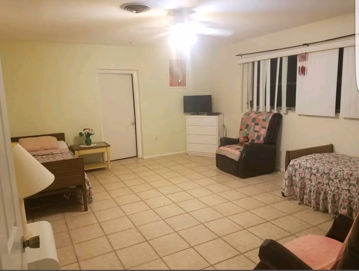 A simple, clean room with two single beds covered in floral bedspreads, a small wooden side table with a vase of flowers, a white dresser with a small TV on top, a cushioned armchair with a patchwork quilt draped over it, tiled floor, and vertical blinds on the window.