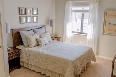 A neatly made bed with a beige patterned bedspread and multiple pillows in a bright bedroom. The room has a window with white curtains letting in natural light, two bedside tables with lamps, and framed artwork on the walls.