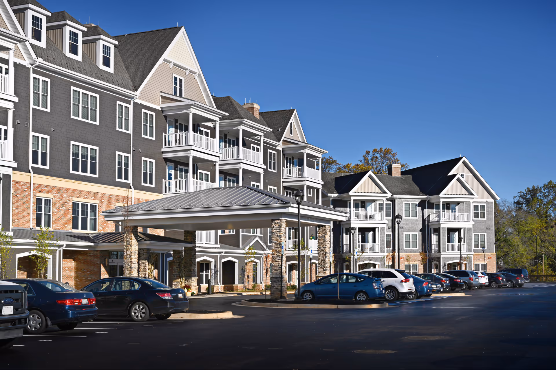 Exterior view of a multi-story senior living community building with balconies, a covered entrance supported by stone pillars, and a parking lot with several parked cars under a clear blue sky.
