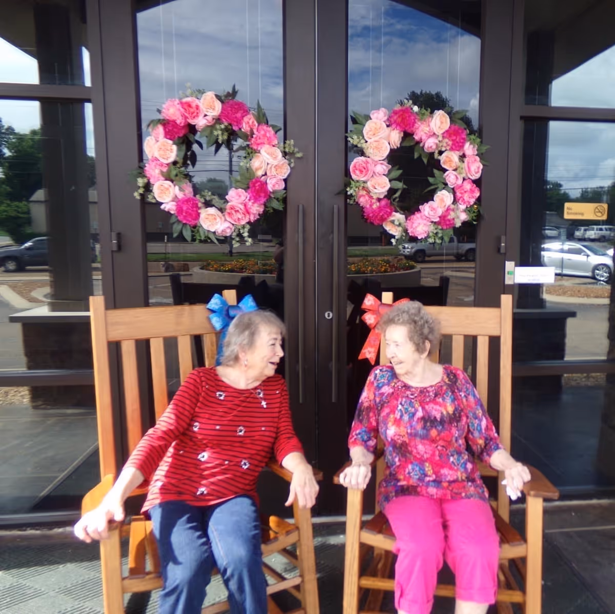 Two older women sit on wooden rocking chairs in front of glass entrance doors decorated with pink floral wreaths.