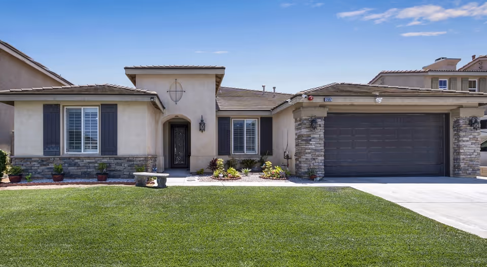 Front exterior view of a single-story residential building with beige stucco walls, stone accents, a dark brown garage door, and a well-maintained green lawn under a clear blue sky.