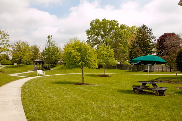 Green outdoor common area with winding paved paths, small trees, a gazebo, benches, and a picnic table with a green umbrella.