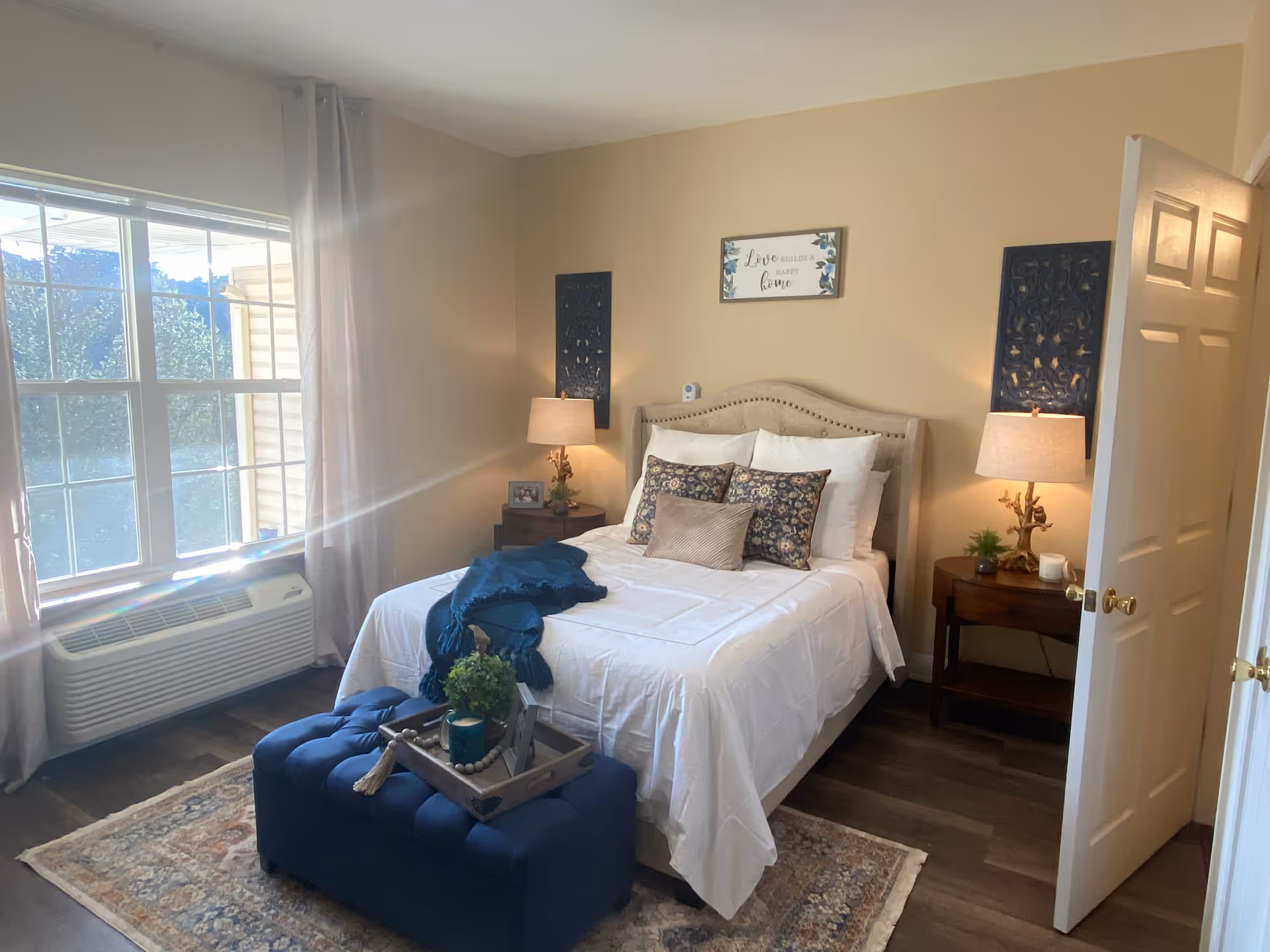 A cozy bedroom with a neatly made bed featuring white bedding and decorative pillows. There are two wooden nightstands on either side of the bed, each with a lamp and small decorative items. A large window with light curtains allows natural light to fill the room. A blue cushioned bench with a tray holding a candle and plant sits at the foot of the bed. The walls are painted beige and decorated with framed art and wall hangings.