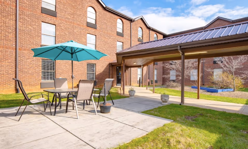 Outdoor patio area at Regency Retirement Village of Morristown featuring a table with four chairs and a teal umbrella, surrounded by a brick building and a covered walkway. The area is sunny with green grass and some potted plants.