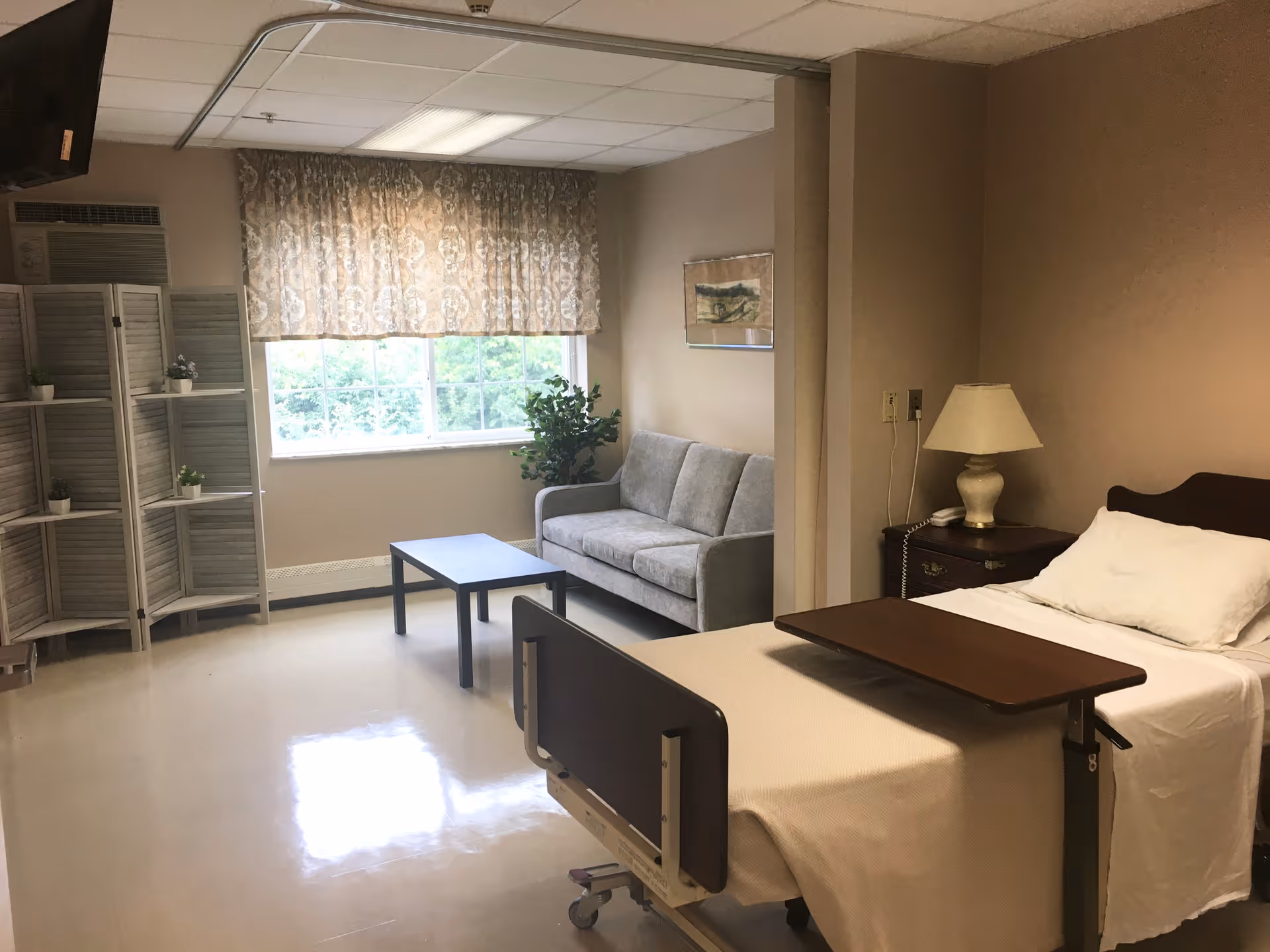 A nursing facility room with a hospital bed, a bedside table with a lamp and phone, a gray couch, a coffee table, a window with patterned curtains, a folding screen with small potted plants, and a wall-mounted TV.