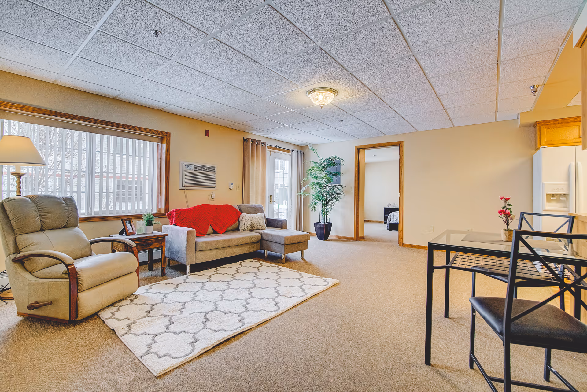 Spacious assisted-living style living room with a gray sofa and red throw, recliner, area rug, dining table, and potted plant.