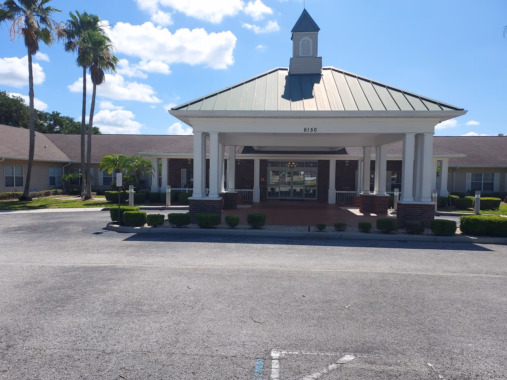 Front exterior view of Hawthorne Inn of Lakeland assisted living and memory care facility with a covered entrance supported by white columns, a green metal roof, and a small cupola on top. The building is surrounded by neatly trimmed bushes and palm trees under a blue sky with scattered clouds.