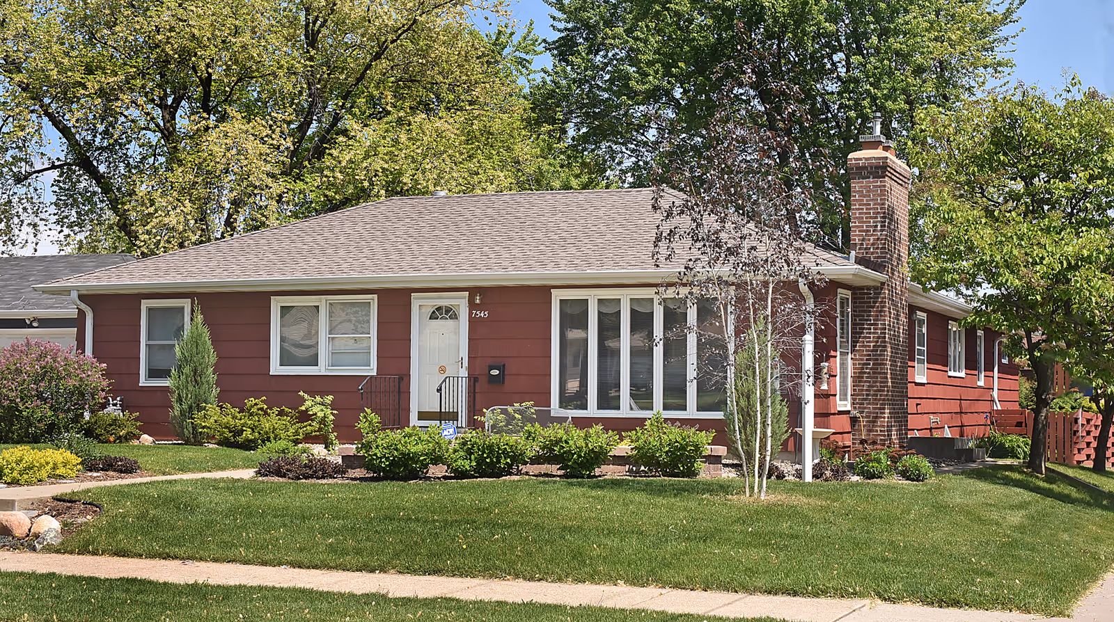 Single-story red house with a gray shingled roof, white front door, and multiple windows. The house is surrounded by green grass, shrubs, and trees under a clear blue sky.