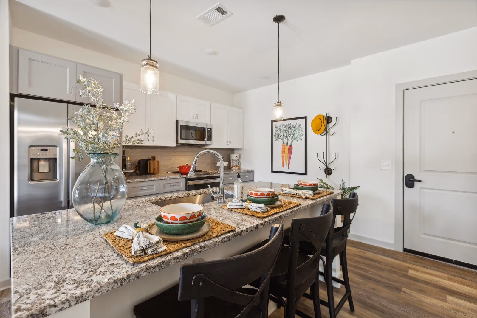 Modern kitchen with a granite countertop island set with three place settings, each with a bowl, plate, and napkin. The kitchen features stainless steel appliances including a refrigerator and microwave, white and gray cabinetry, two pendant lights hanging above the island, a large glass vase with leafy branches on the counter, and a framed carrot artwork on the wall near the entrance door.