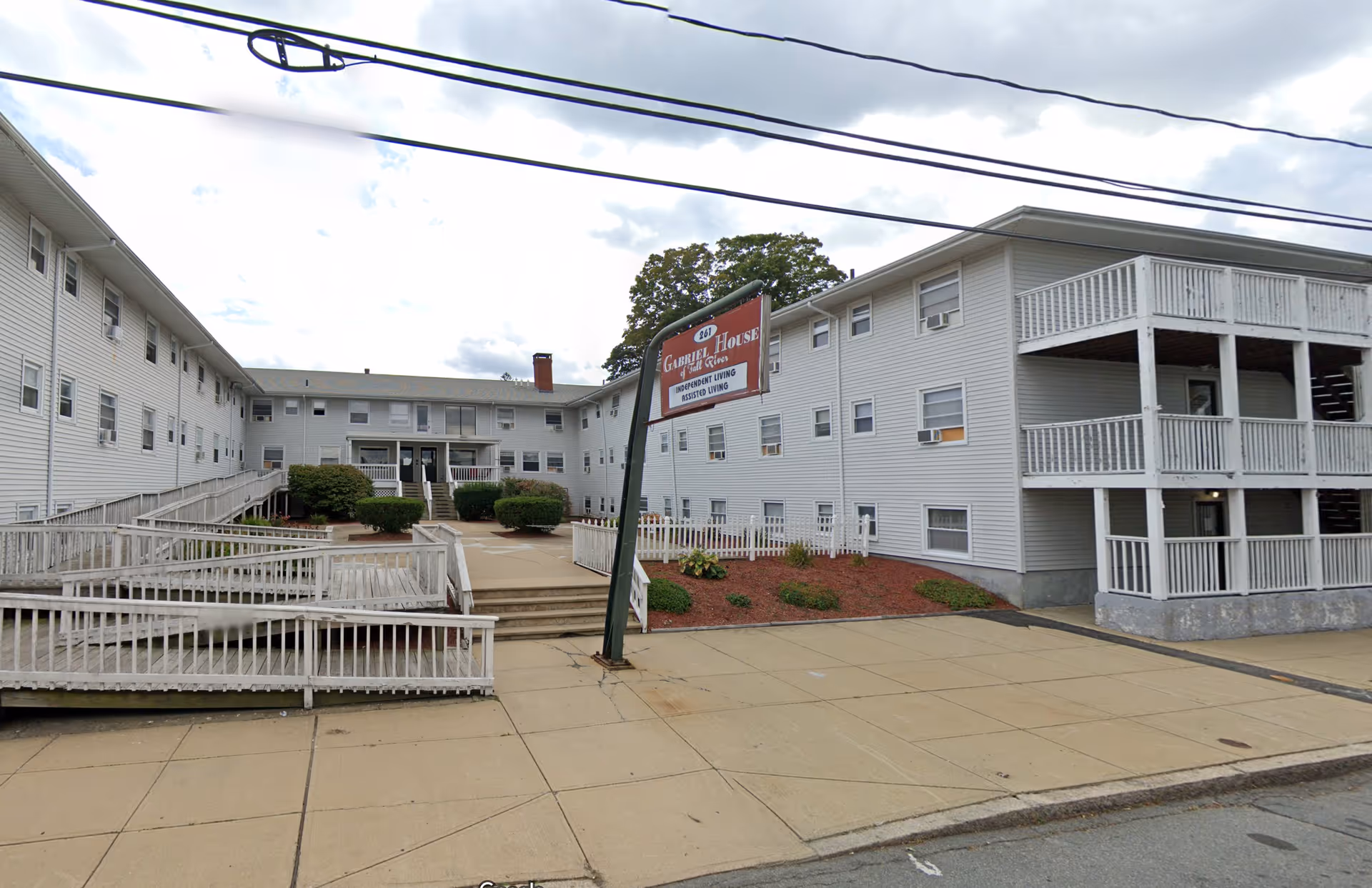 Front exterior of a multi-story white assisted living building with ramps, balconies, landscaping, and a sign for Gabriel House.