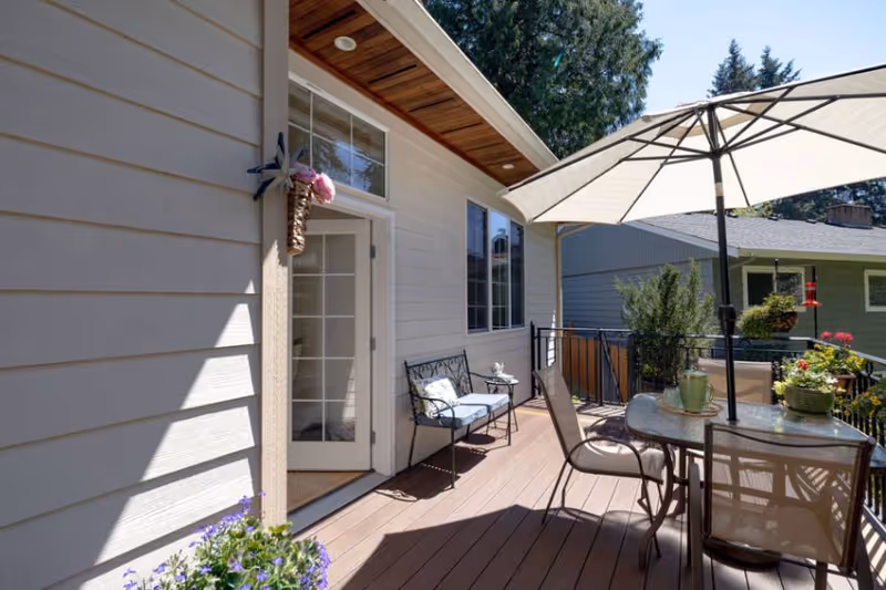 A sunny residential deck with a patio table and umbrella, chairs, potted plants, and a glass door leading into the house.