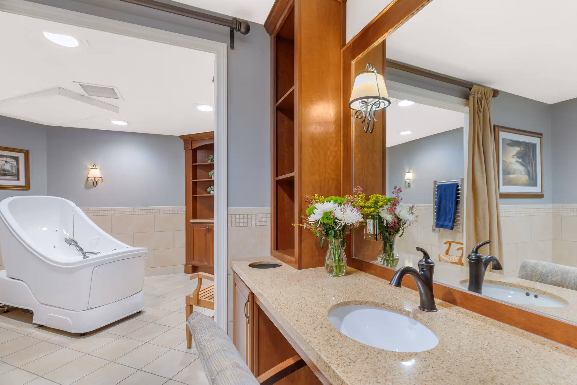 A clean and well-lit bathroom area in a senior living facility featuring a beige countertop with a sink and a bronze faucet. A vase with colorful flowers is placed on the counter. There is a large mirror above the sink with a wall-mounted light fixture. In the background, a white therapeutic bath chair is visible, along with a wooden chair, a towel rack with a blue towel, and framed artwork on the walls. The walls are painted light blue with beige tile halfway up.