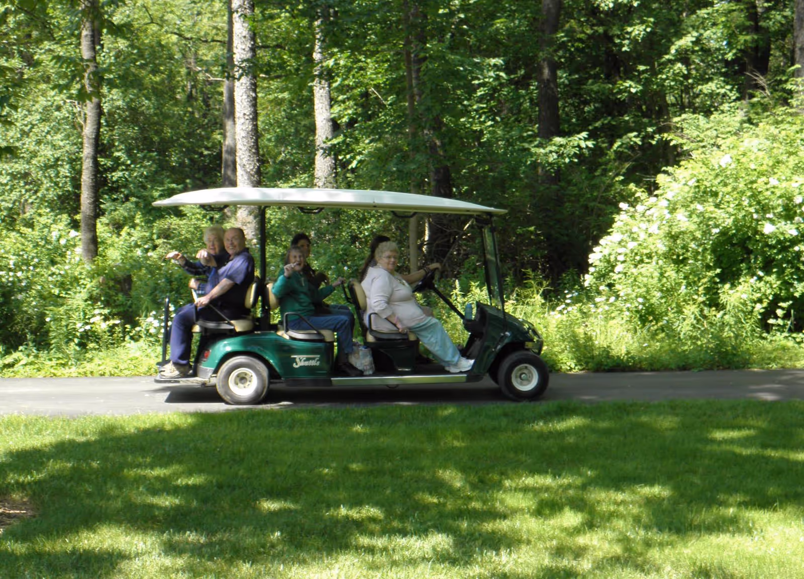 A group of elderly people riding together in a green golf cart on a paved path surrounded by lush green trees and grass.