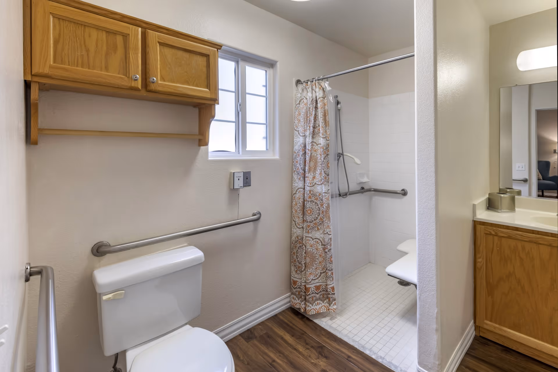 A bathroom with a white toilet, wooden cabinet mounted on the wall, a window, and a walk-in shower with a patterned shower curtain and grab bars. The shower has a built-in seat and handheld showerhead. There is a wooden vanity with a sink and a mirror on the right side.