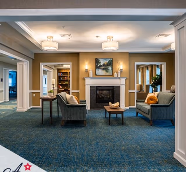 A cozy common room with sofas and chairs arranged around a central fireplace, coffee table, artwork, and overhead lighting.