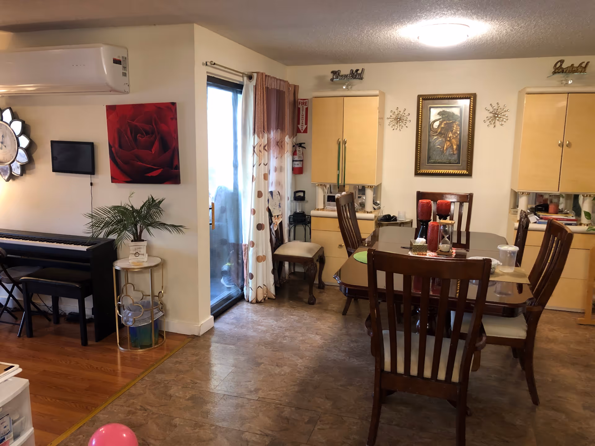 Interior view of a dining area with a wooden dining table and six chairs. The table has decorative items including red candles and a tissue box. Behind the table, there are two light wood cabinets mounted on the wall with decorative wall art and two metallic wall decorations. To the left, there is a sliding glass door with curtains, a small round side table with a potted plant, a wall-mounted air conditioner, a large wall clock, and a digital device mounted on the wall. A piano with a bench is also visible on the left side of the image.