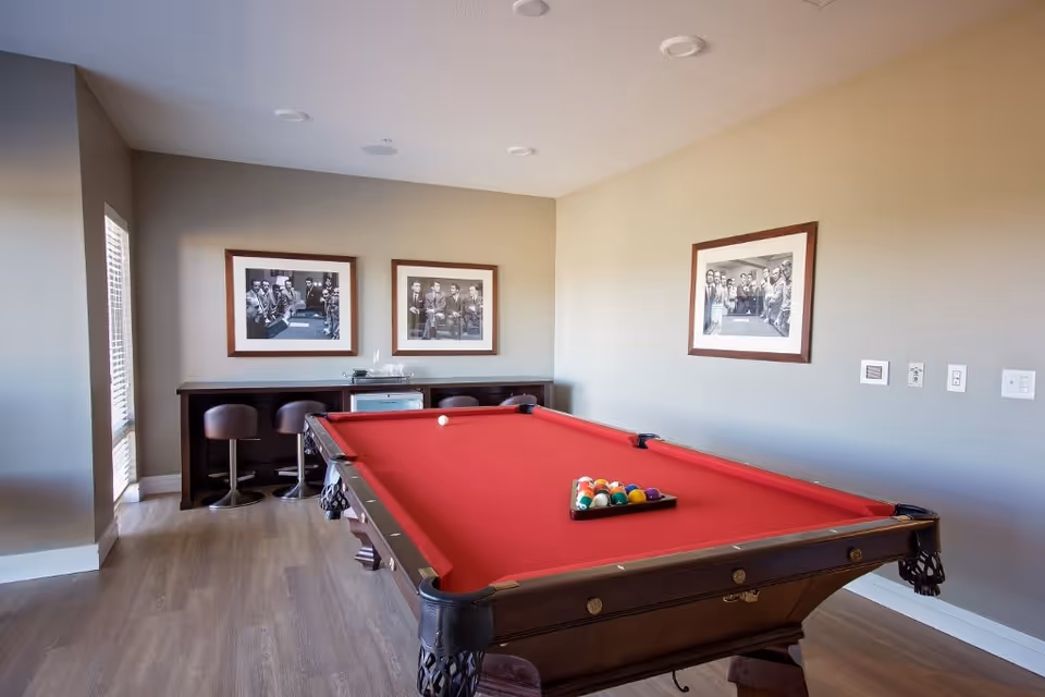 A recreational room with a red felt pool table in the center, a set of pool balls arranged on the table, three bar stools along a counter against the wall, and three framed black and white photographs hanging on the walls.