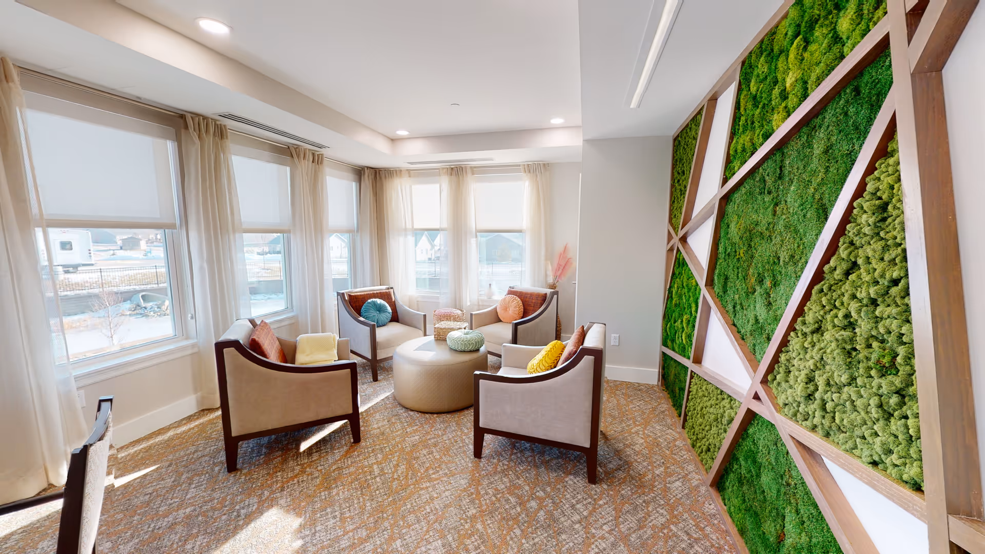 A bright and cozy sitting area with four beige armchairs arranged around a round ottoman. The chairs have colorful cushions in yellow, orange, and teal. Large windows with sheer curtains let in natural light. One wall features a decorative green moss panel with wooden framing, adding a natural element to the room.