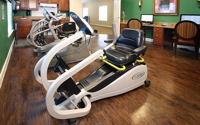 Indoor exercise room with wooden flooring featuring a NuStep recumbent cross trainer and other cardio equipment. The room has green walls, wooden cabinets, and a desk area with two chairs and computer monitors.