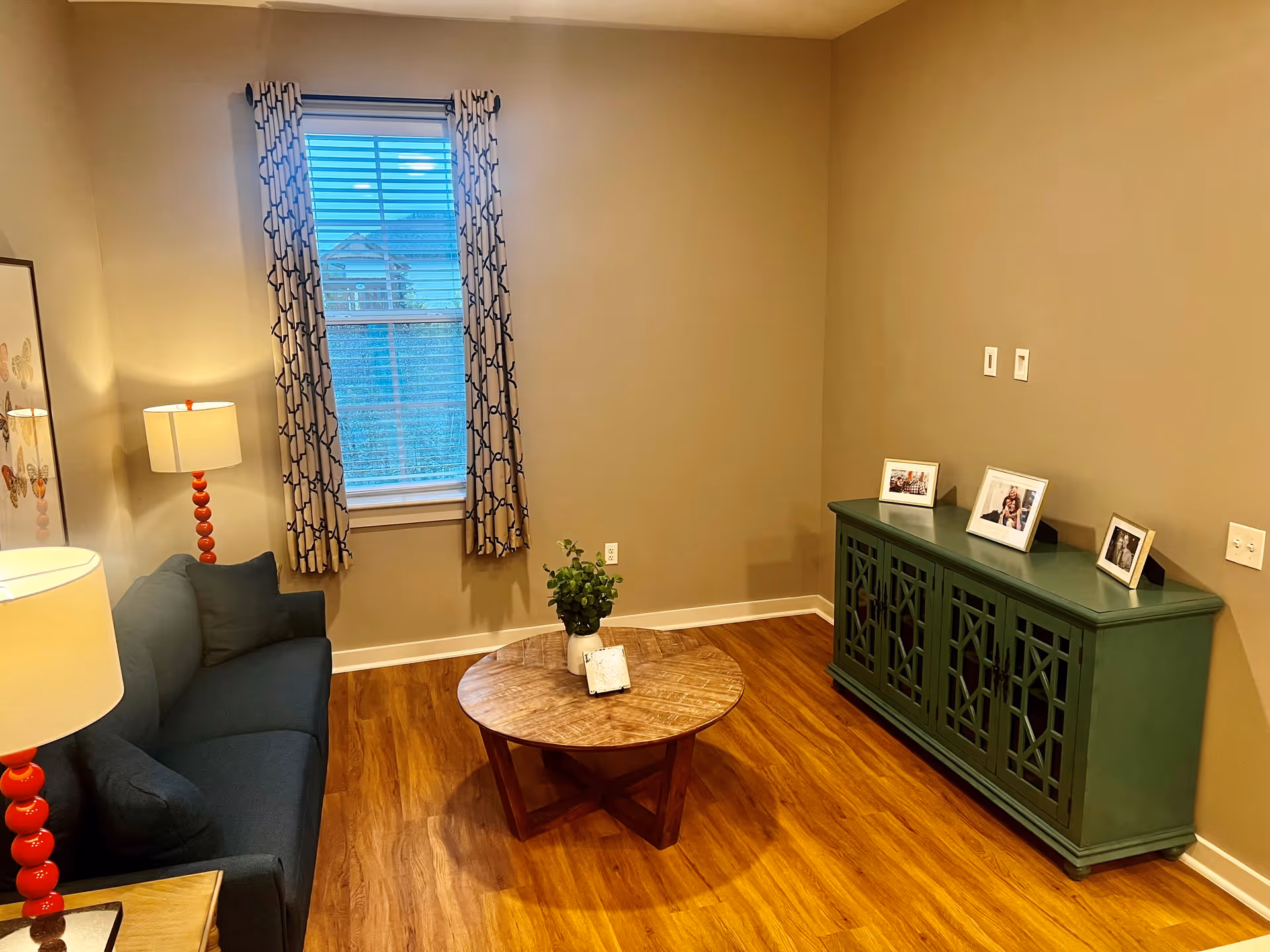 Small living room with a navy sofa, round wooden coffee table, green console cabinet, and a window with patterned curtains.