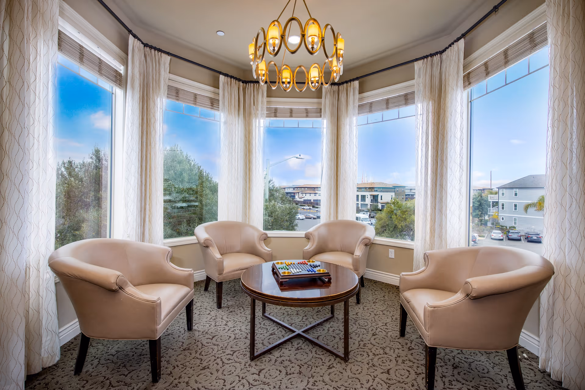 A cozy seating area with four beige armchairs arranged around a round wooden table with a board game on it. The space is surrounded by large windows with white curtains, offering a view of trees, buildings, and parked cars outside. A modern chandelier hangs from the ceiling above the table.