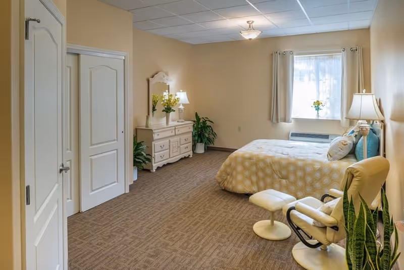 A cozy bedroom with a bed covered in a yellow patterned comforter, a white dresser with a mirror and lamp, a comfortable armchair with an ottoman, potted plants, and a window with curtains letting in natural light.