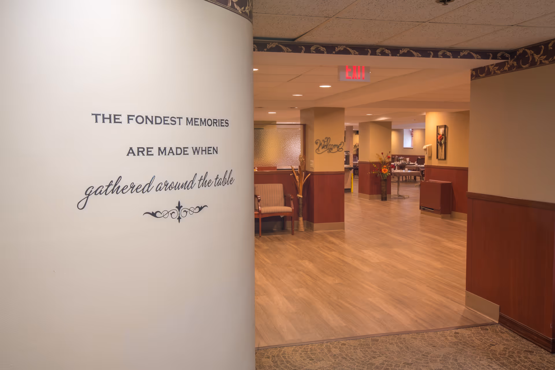 Interior view of a nursing facility hallway opening into a dining area with a wall quote reading "The fondest memories are made when gathered around the table."