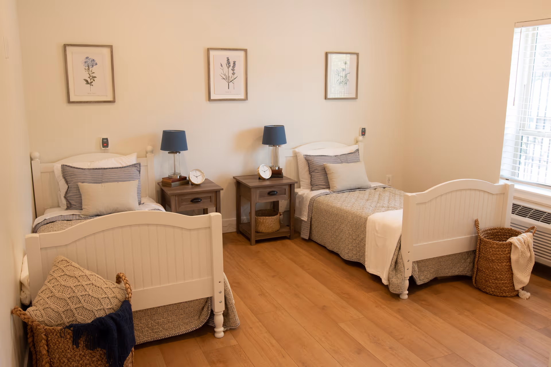 A cozy bedroom with two single beds featuring white wooden frames and beige bedding. Each bed has a small pillow and a larger pillow with striped pillowcases. Between the beds are two wooden nightstands, each with a blue lamp and a small clock. Above the beds are three framed botanical prints. The room has light-colored walls and wooden flooring. There is a window with white blinds on the right side and a woven basket with a blanket near each bed.