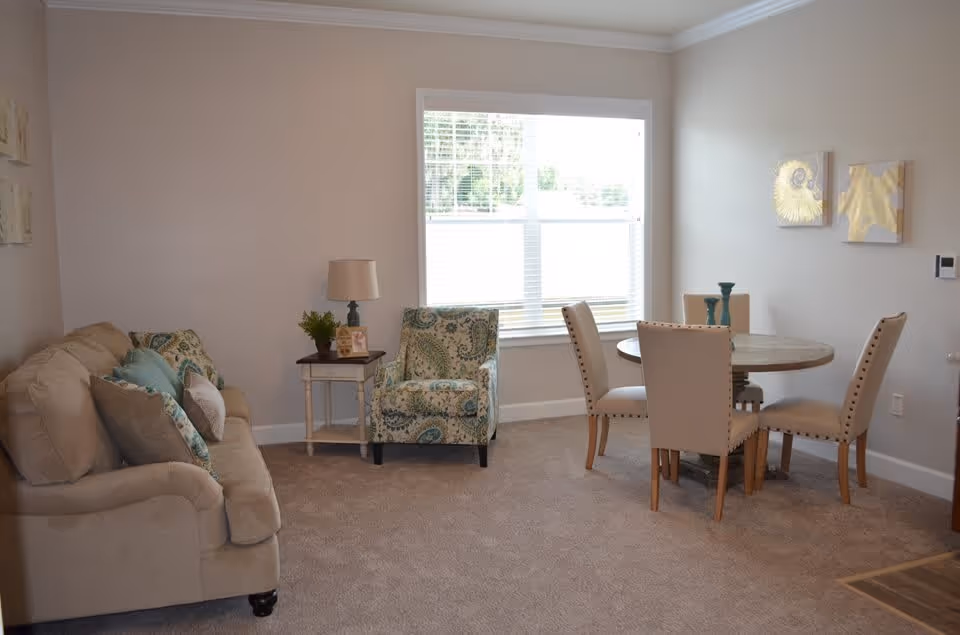A cozy living and dining area with a beige sofa adorned with multiple cushions, a patterned armchair, a small side table with a lamp and plant, and a round dining table surrounded by four beige upholstered chairs. The room has a large window with blinds, light-colored walls, and carpeted flooring.