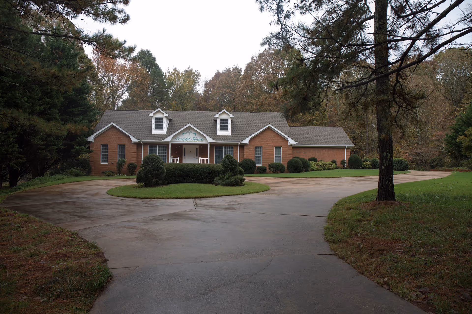 A single-story brick building with a gray roof and white trim, surrounded by trees and greenery. The building has a front porch with two white rocking chairs and a sign above the entrance that reads 'Peaceful Hearts'. A curved driveway leads up to the building, with a grassy island in the center.
