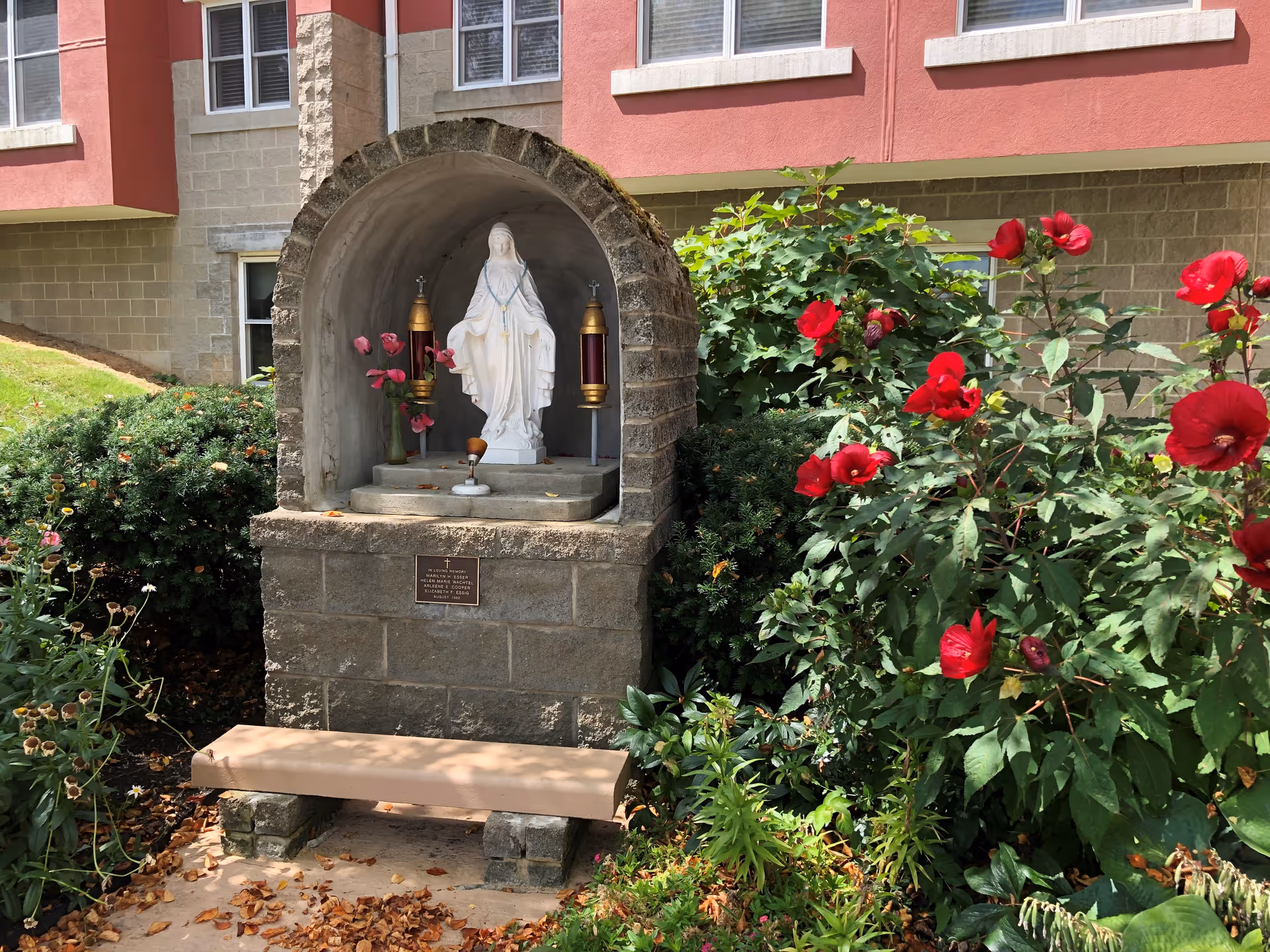 Outdoor shrine with a white statue of the Virgin Mary inside a stone alcove, flanked by two lanterns and a vase with pink flowers. The shrine is surrounded by green bushes and red flowers, with a small bench in front. The background shows part of a building with red and beige walls and windows.